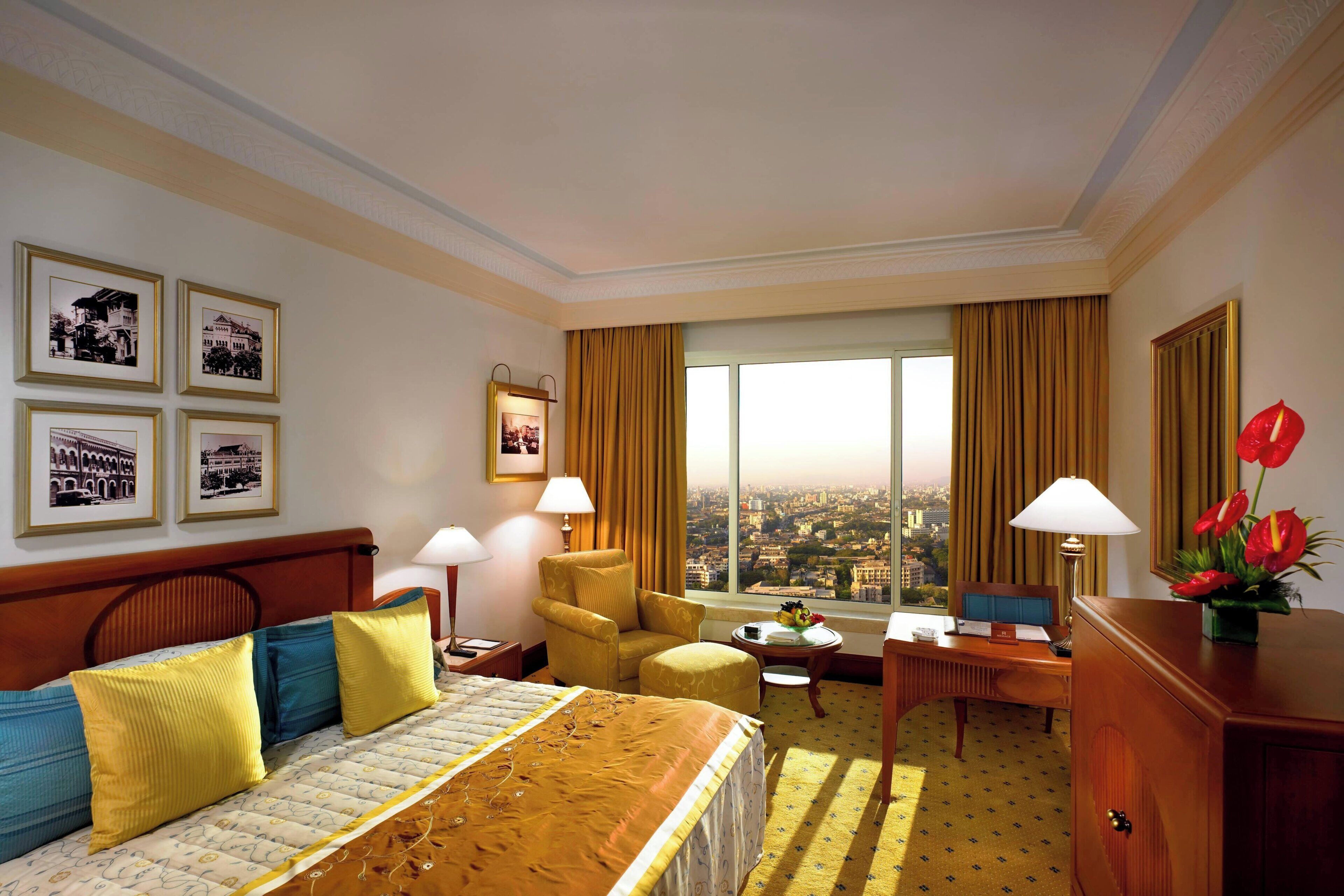 A hotel room with a bed, a desk, a chair, a view of the city, and framed pictures on the wall.