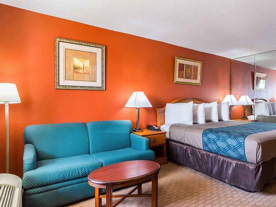 A hotel room with a teal sofa, a wooden table, a bed with a patterned bedspread, a nightstand with a lamp, and two framed pictures on the wall.