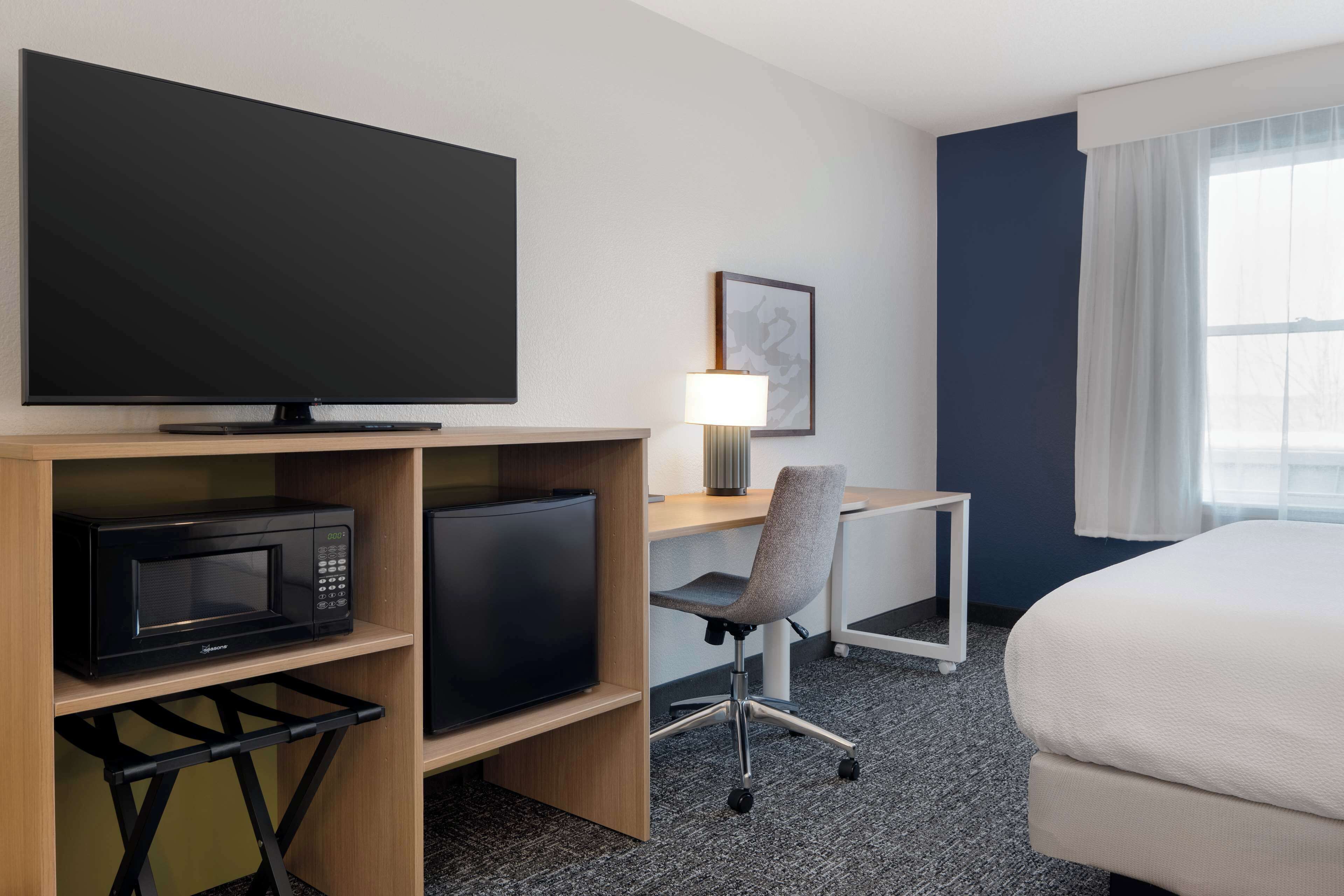 A hotel room with a flat-screen TV, a microwave, a desk with a chair, a lamp, and a bed.