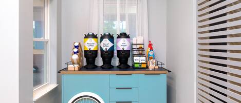 Um carrinho azul-esverdeado com três dosadores de bebidas identificados como 'Café Comum', 'Café Descafeinado' e 'Água Quente', e uma embalagem de sabonete líquido.