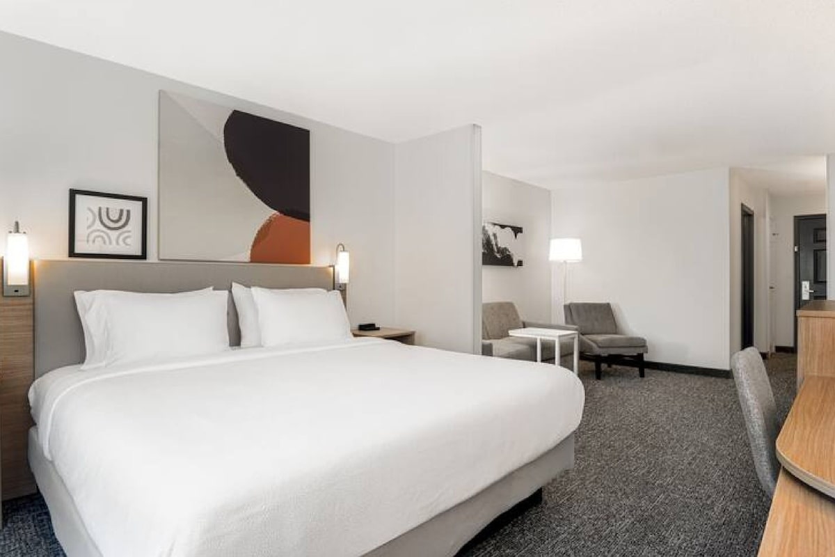 A hotel room with a large bed, a desk, a chair, and a small table. There are wall art pieces and a door visible.