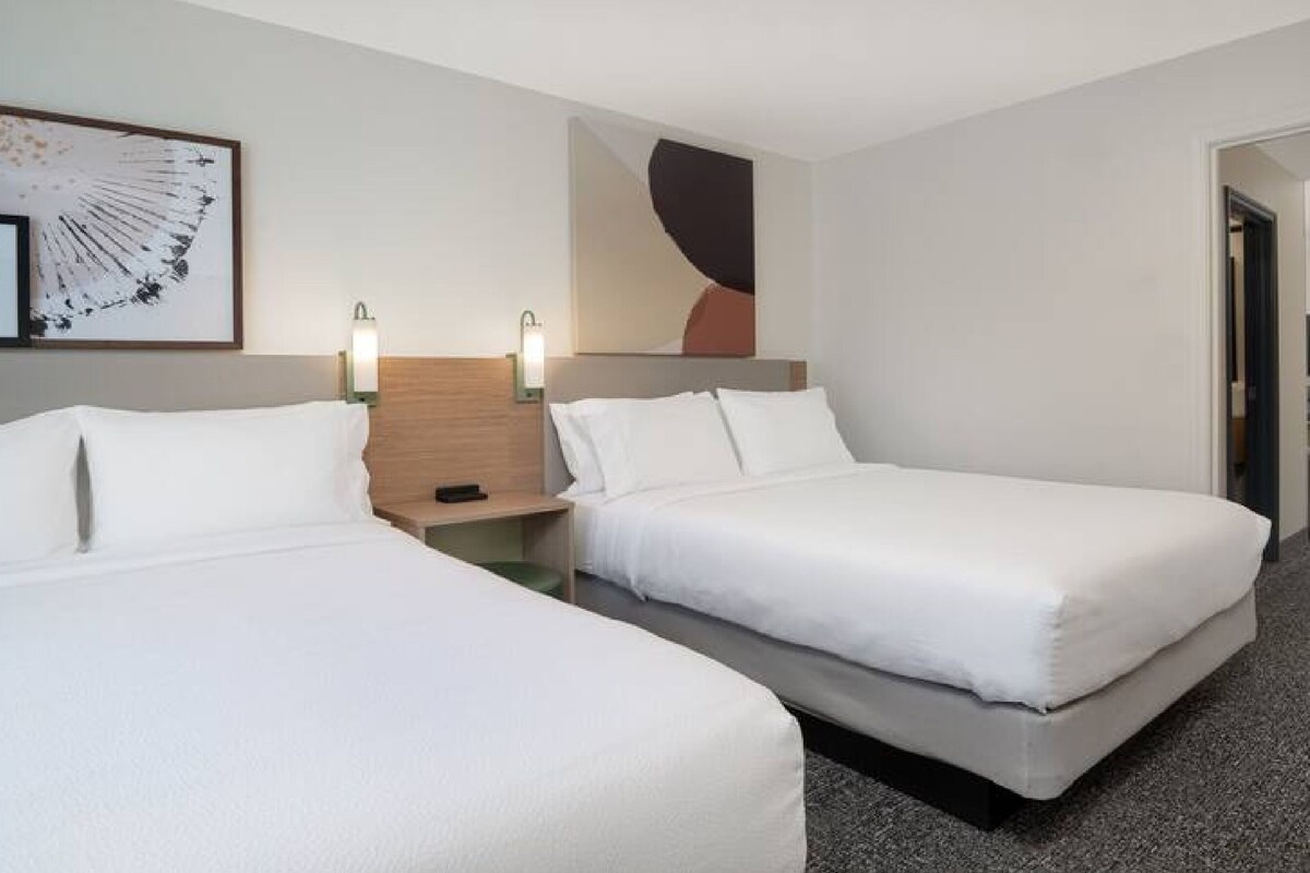 A hotel room with two single beds, a wooden headboard, a wall-mounted light, and abstract art.
