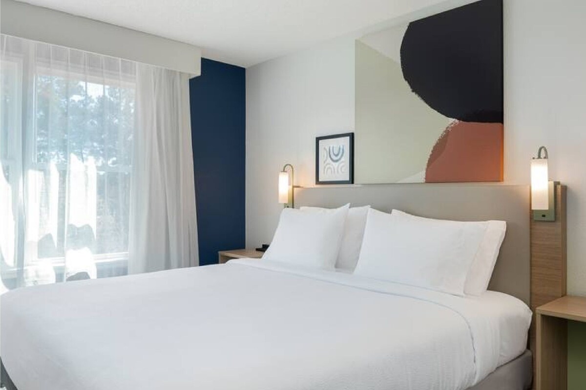 A hotel room with a large bed, a nightstand, a wall-mounted lamp, and a window with sheer curtains.