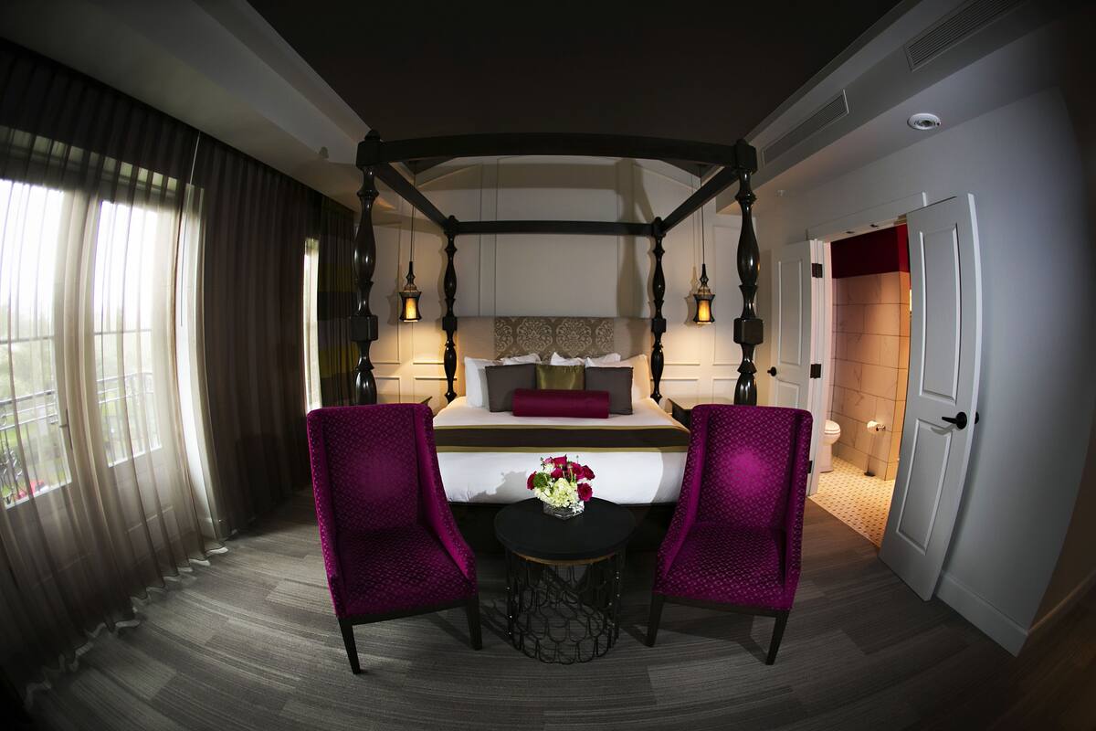 Hotelkamer met hemelbed, twee paarse fauteuils, een klein rond tafeltje met een bloevas en uitzicht buiten door dunne gordijnen.