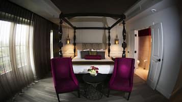 A hotel room with a canopy bed, two purple armchairs, a small round table with a vase of flowers, and a view of the outside through sheer curtains.