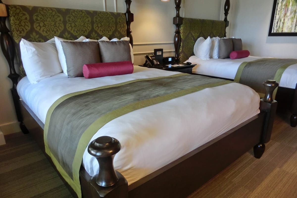 Een tweepersoonsbed met een houten hoofdeinde en voeteneinde, een nachtkastje met een telefoon, en een bedtafel met een lamp.