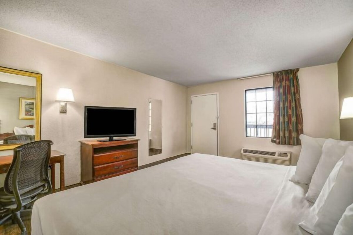 A hotel room with a bed, a desk with a chair, a television, a window with curtains, and a door.
