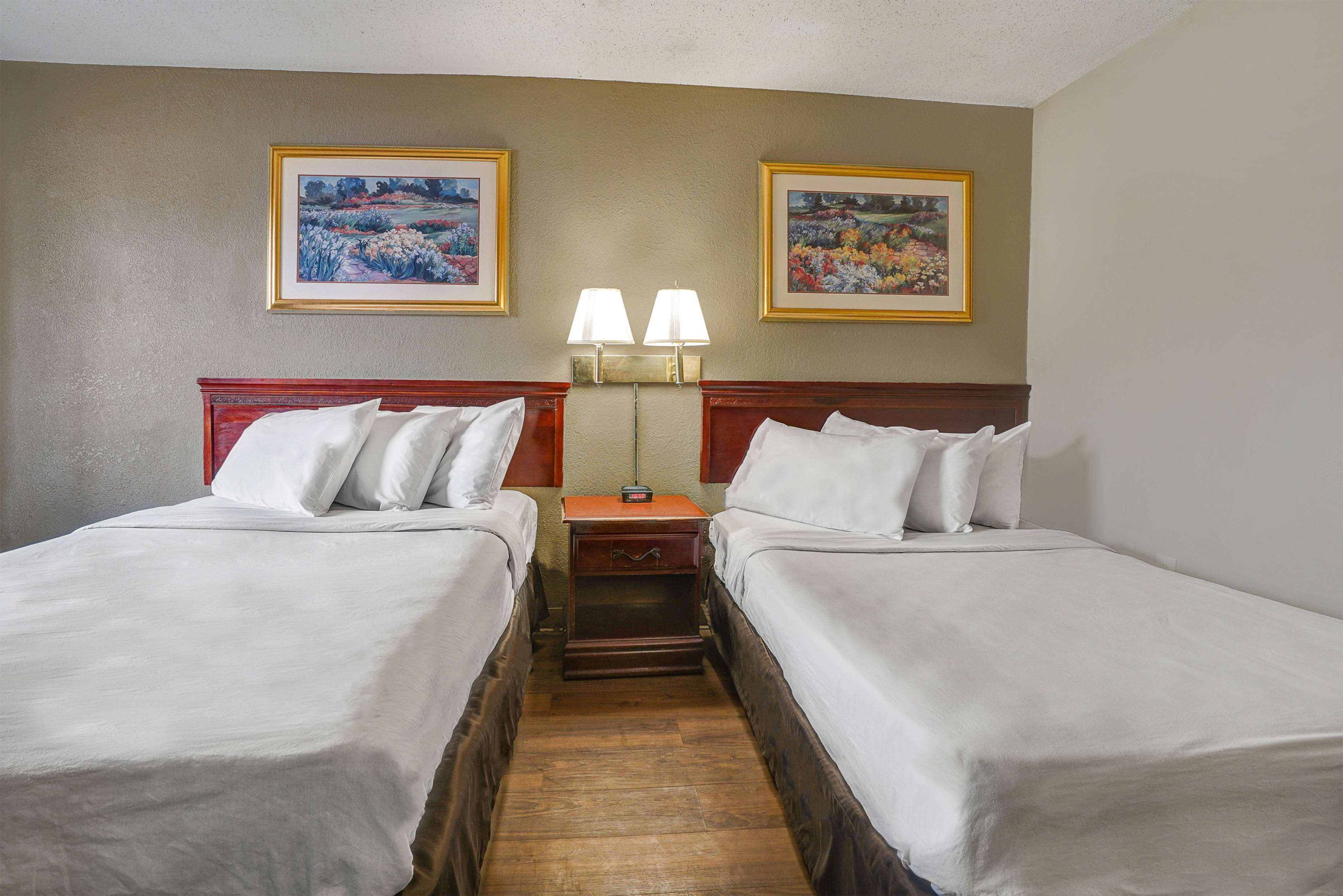 Habitación de hotel con dos camas, una mesita de noche con lámparas y dos cuadros enmarcados con motivos florales en la pared.