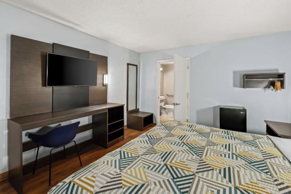 A modern hotel room with a large bed, a desk with a chair, a flat-screen TV mounted on the wall, a small heater, and a door leading to a bathroom.