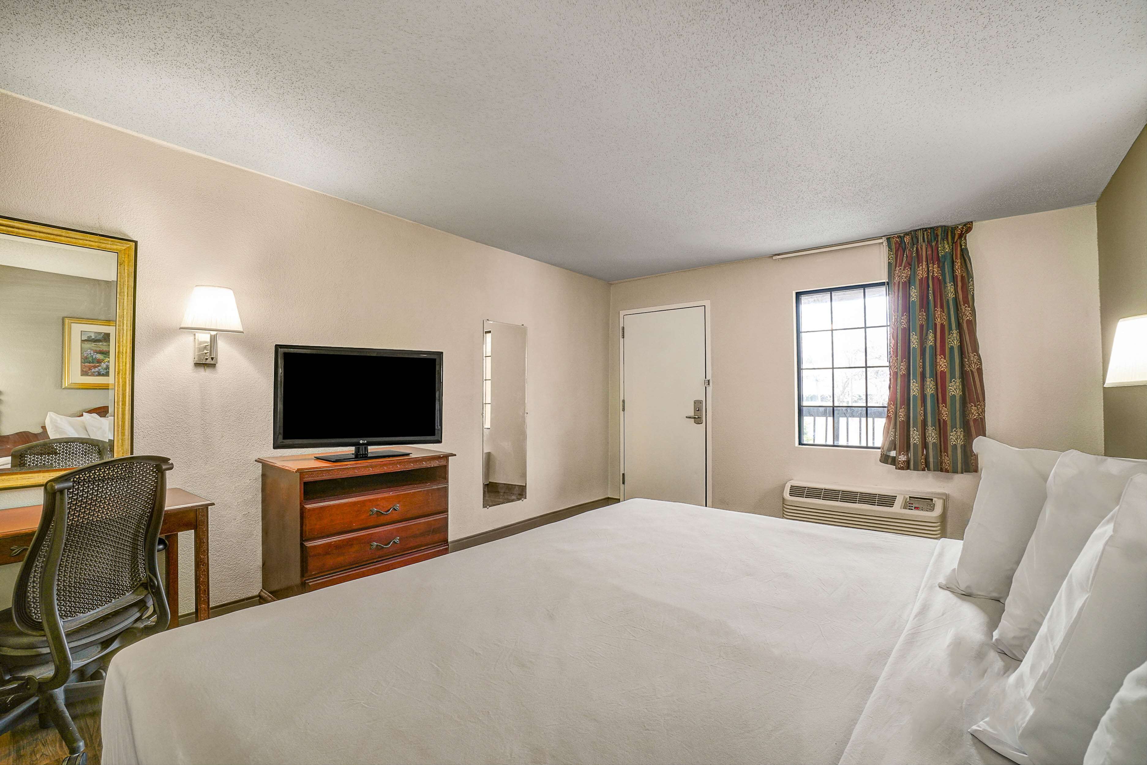 Habitación de hotel con cama, escritorio con silla, televisor, espejo, ventana con cortinas y calentador de pared.