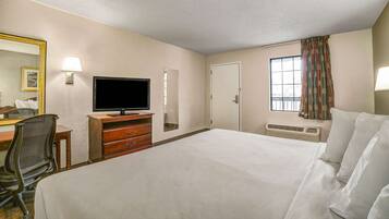 Habitación de hotel con cama, escritorio con silla, televisor, espejo, ventana con cortinas y calentador de pared.
