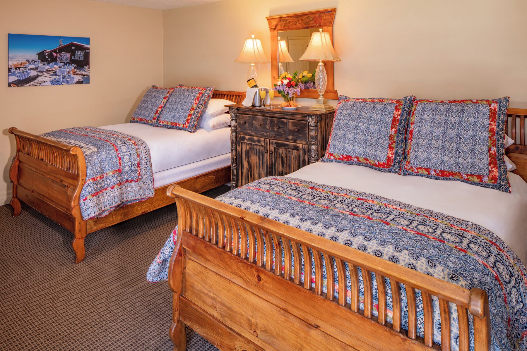 A room with two wooden beds, each with patterned bedding and pillows. There is a wooden dresser with a mirror above it, and a painting of a building on the wall.