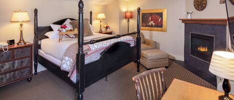A four-poster bed with a wooden headboard, a bedside table with a lamp, a small table with a lamp, a fireplace, and a painting on the wall.