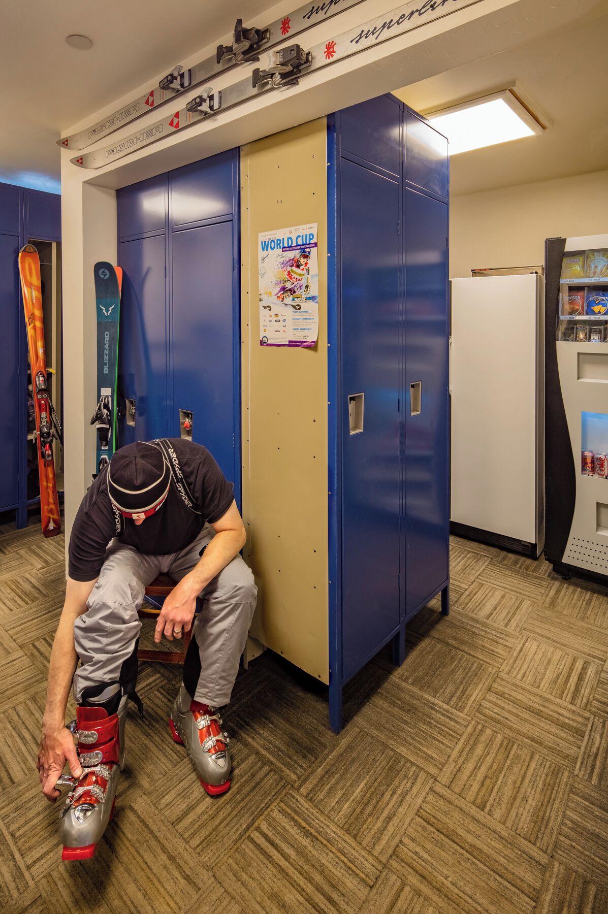 A locker room with blue lockers, a poster on the wall, and a vending machine.