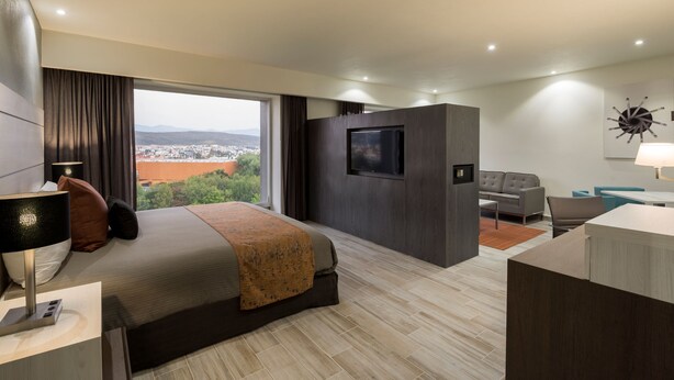 A modern hotel room with a large bed, a TV, a desk, and a view of a cityscape. - Real Inn San Luis Potosí (San Luis Potosi)