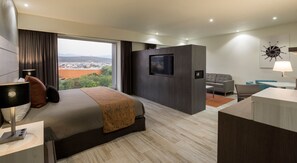 A modern hotel room with a large bed, a TV, a desk, and a view of a cityscape.