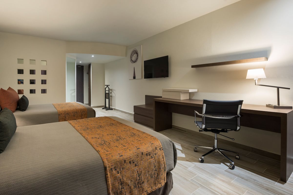 A hotel room with two beds, a desk, a chair, a TV, and a lamp.