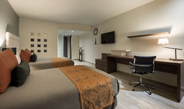 A hotel room with two beds, a desk, a chair, a TV, and a lamp.
