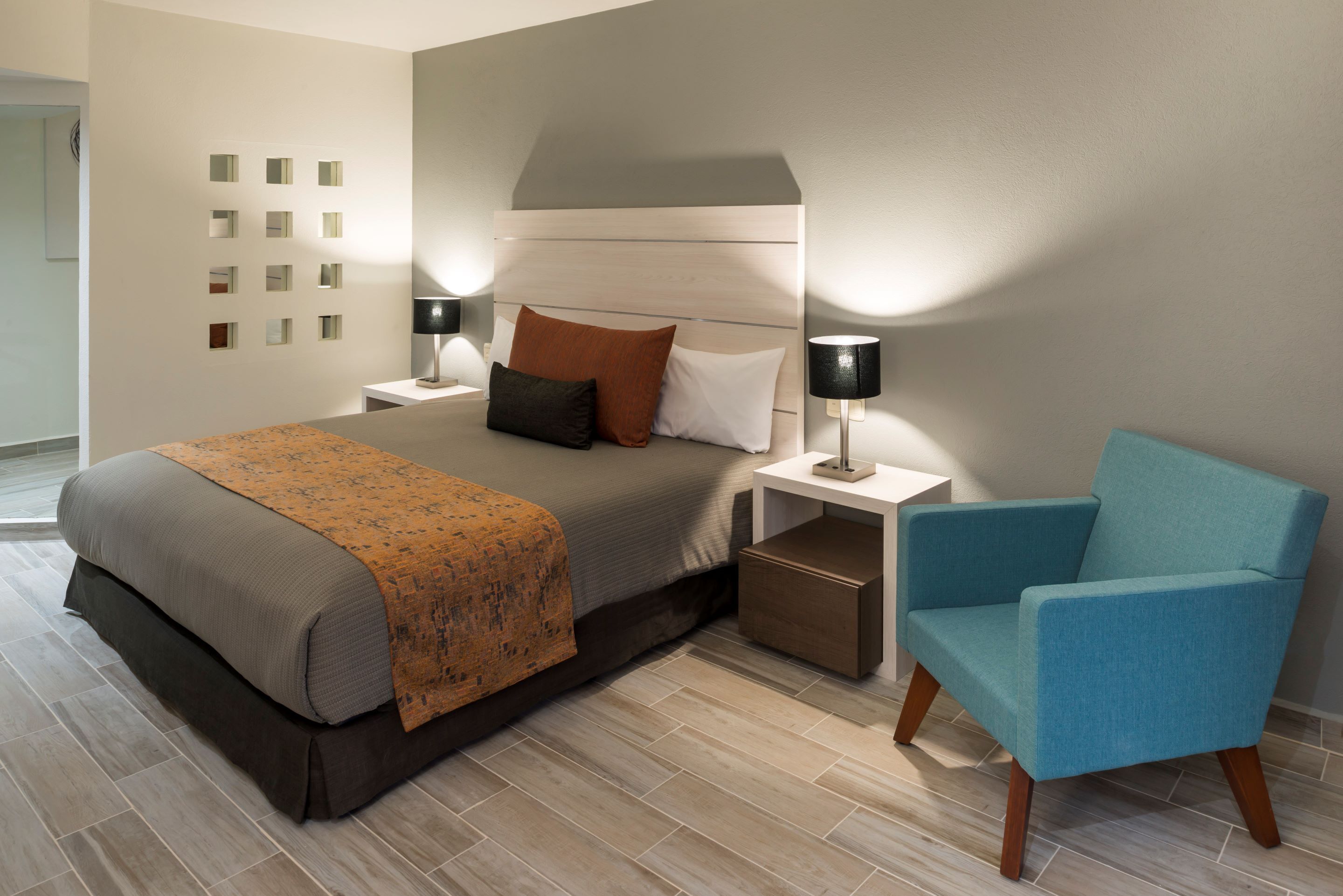 A modern bedroom with a bed, nightstands, a blue armchair, and a wall with small square mirrors.