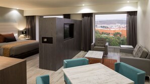A modern hotel room with a bed, a TV, a dining area with a table and chairs, and a view of the cityscape. - Real Inn San Luis Potosí (San Luis Potosi)