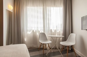 Quarto de hotel com cama, duas cadeiras brancas, uma mesinha redonda e uma janela grande com cortinas leves.
