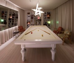 Uma mesa de bilhar com um candeeiro em forma de estrela acima, rodeada por estantes de livros e duas poltronas.