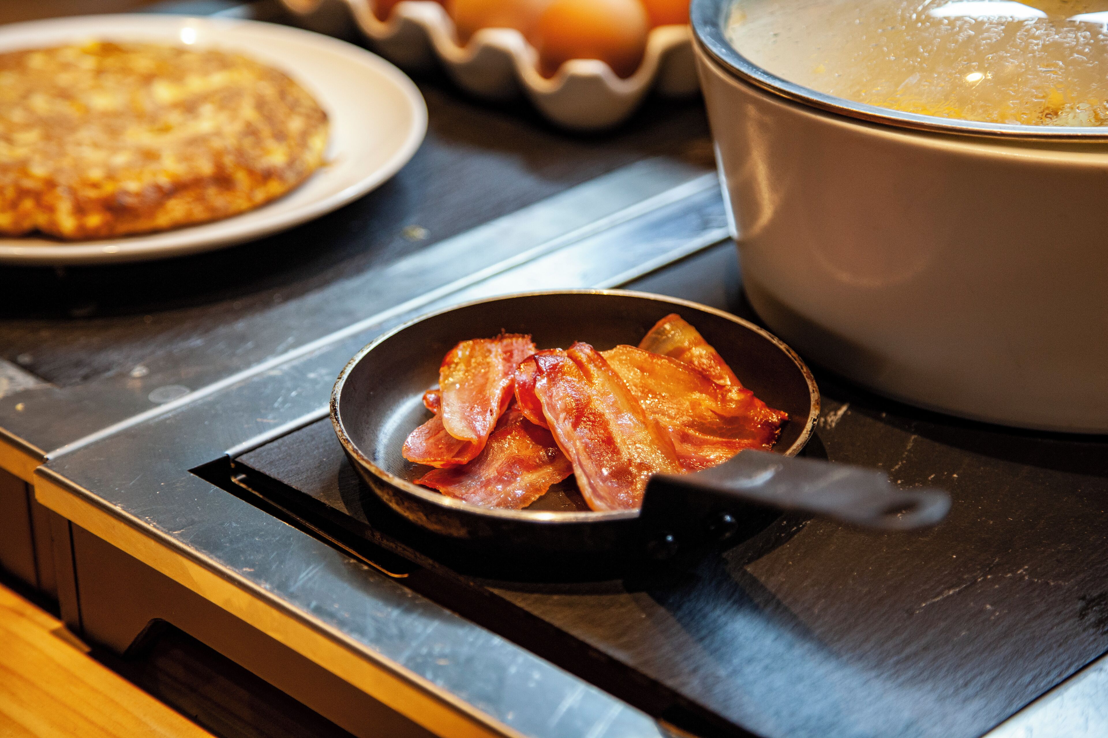 Bacon sizzling in a pan on a stovetop, with a pot and a plate of food in the background.