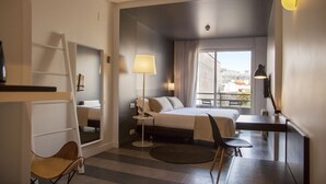 A hotel room with a bed, desk, chair, lamp, and a view of the city through the window.