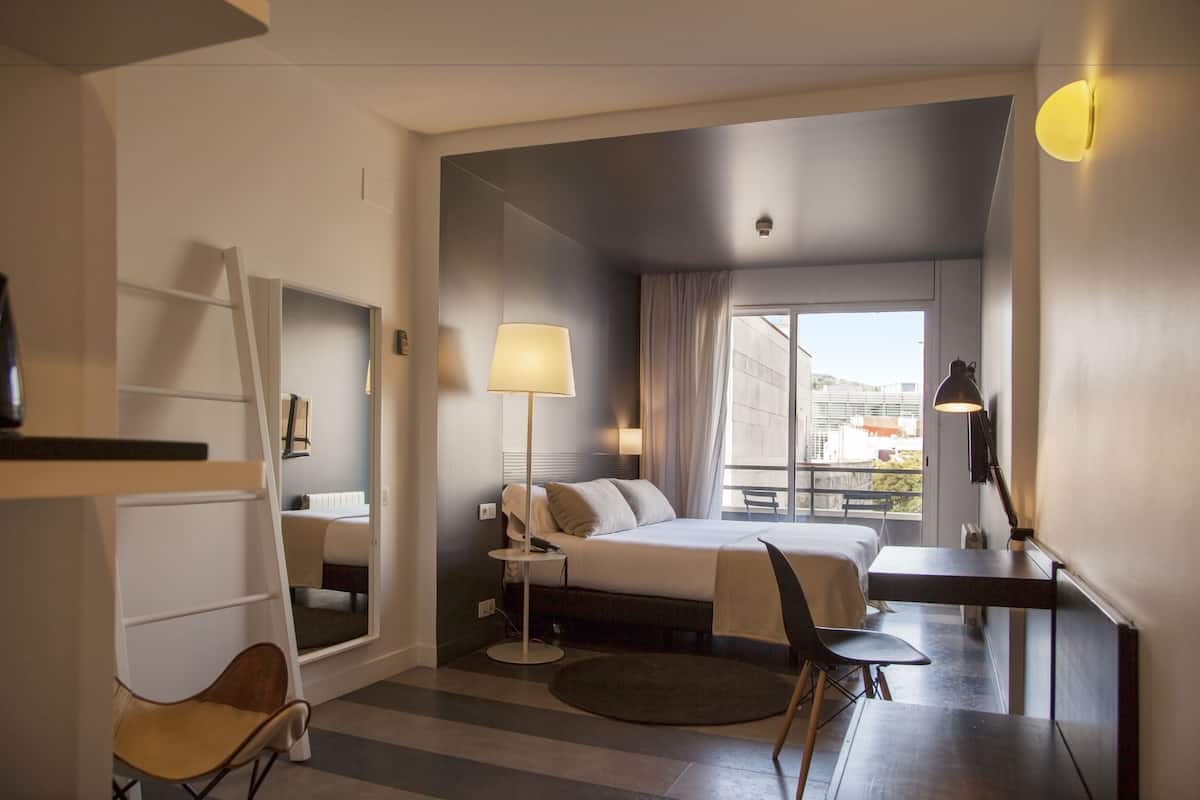 Habitación de hotel con cama, escritorio, silla, lámpara y vista a la ciudad por la ventana.