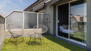 A balcony with two wireframe chairs and a small table, a sliding glass door leading to a bedroom, and a grey exterior wall.