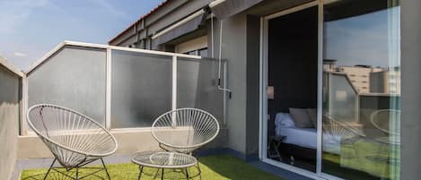 A balcony with two wireframe chairs and a small table, a sliding glass door leading to a bedroom, and a grey exterior wall.