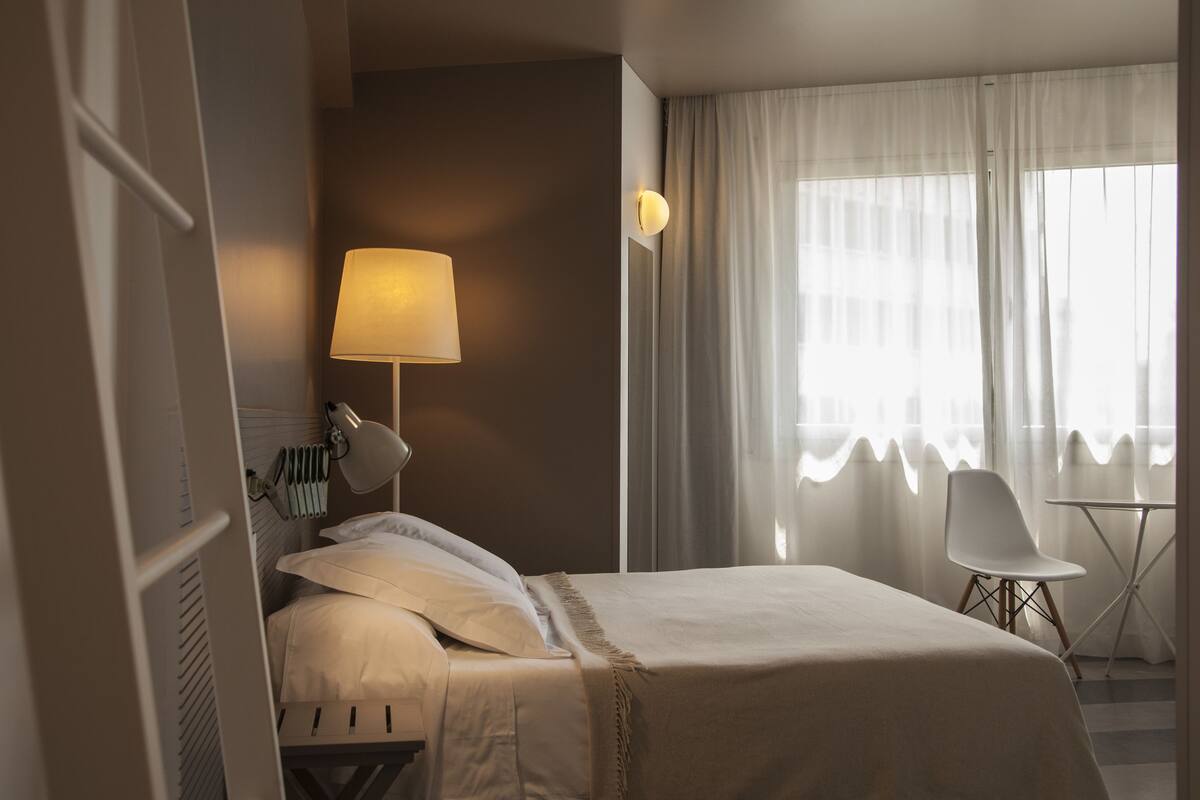 Una habitación de hotel con cama, lámpara de noche, una silla, una mesita y una ventana con cortinas.