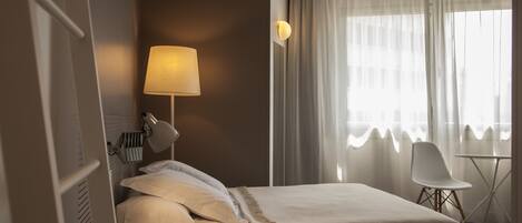 Una habitación de hotel con cama, lámpara de noche, una silla, una mesita y una ventana con cortinas.