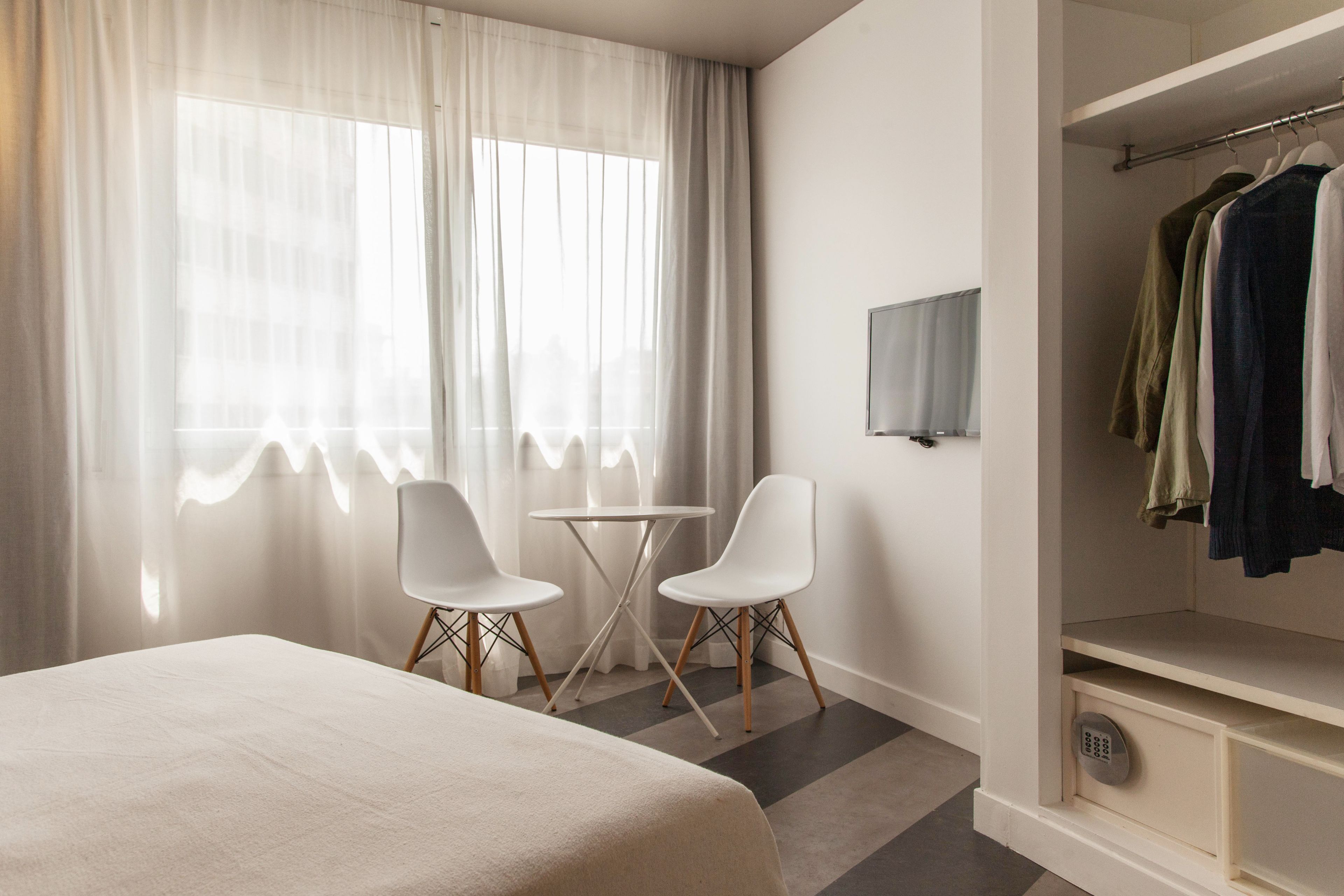 A hotel room with a bed, two white chairs, a small table, a TV, and a wardrobe with clothes.