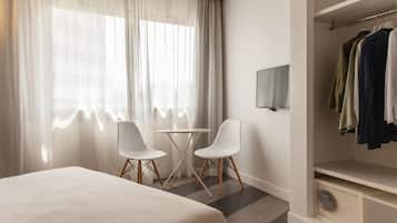 A hotel room with a bed, two white chairs, a small table, a TV, and a wardrobe with clothes.