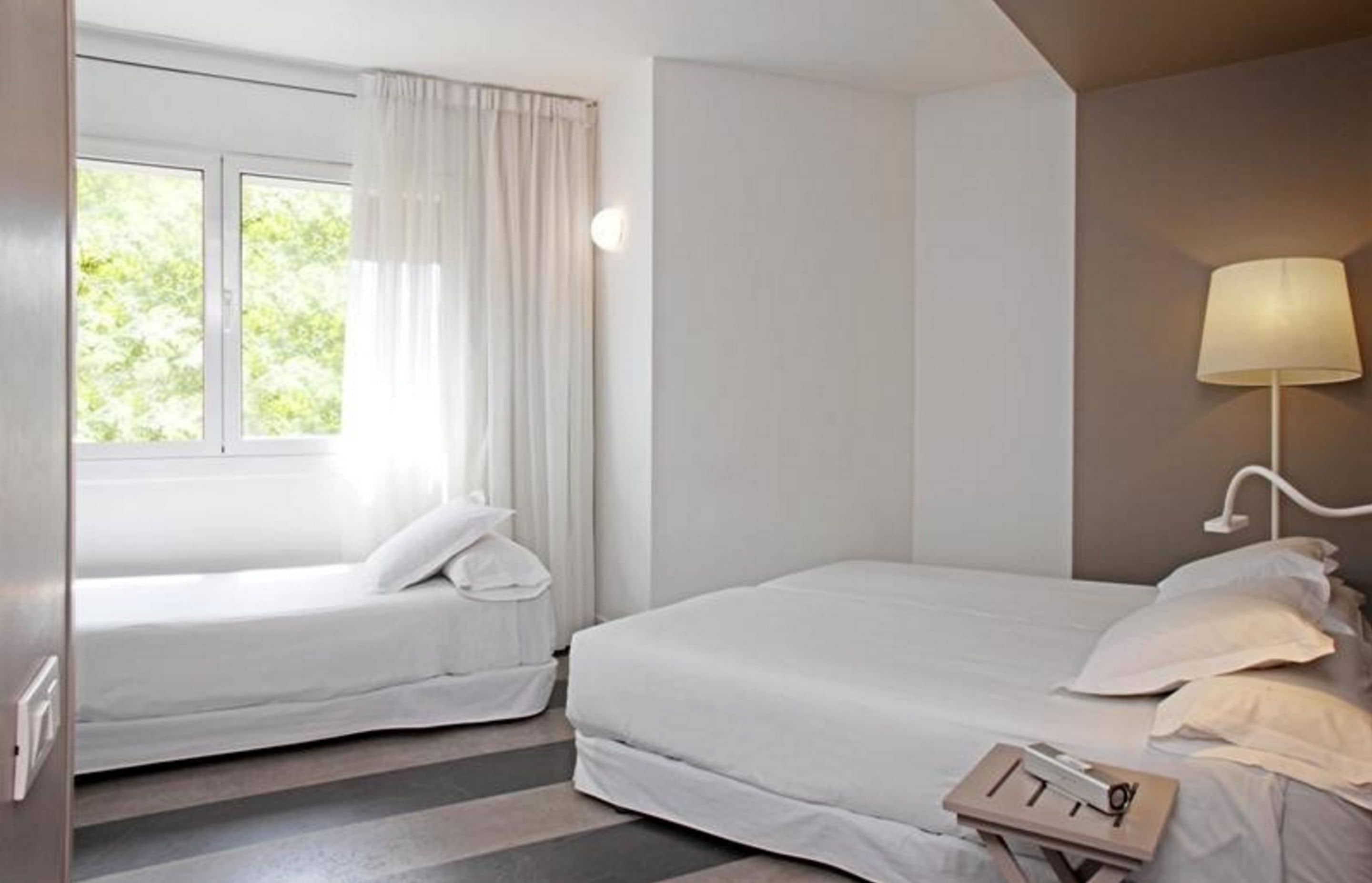Ein Hotelzimmer mit zwei Einzelbetten, einem Fenster mit Vorhängen, einem Nachttisch mit Lampe und Blick auf Grünfläche.
