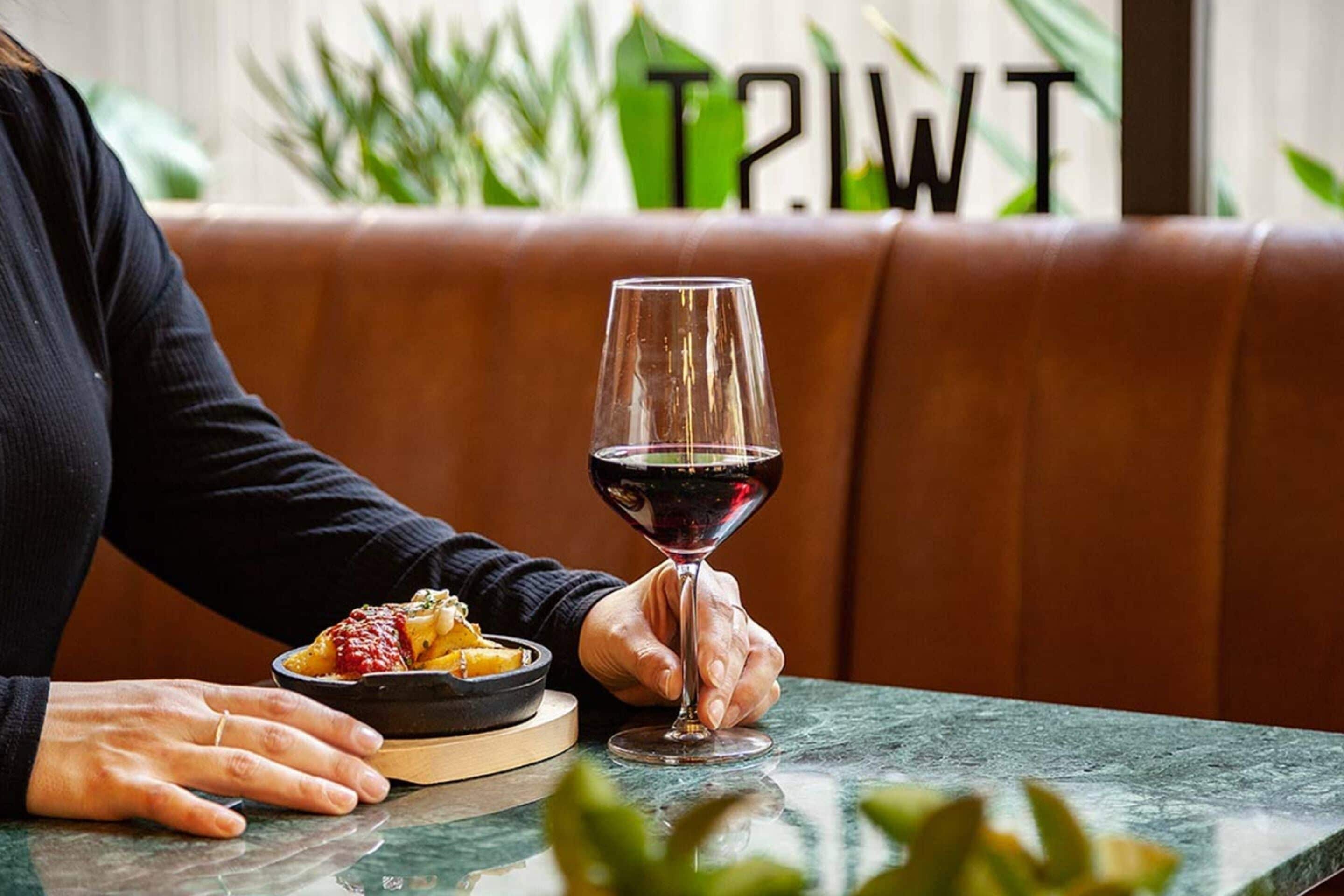 Una persona sentada a una mesa con una copa de vino tinto y un plato de comida.