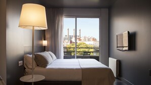 A hotel room with a bed, a bedside lamp, a TV, and a view of a cityscape through a large window. - Sixties Ramblas (Barcelona)