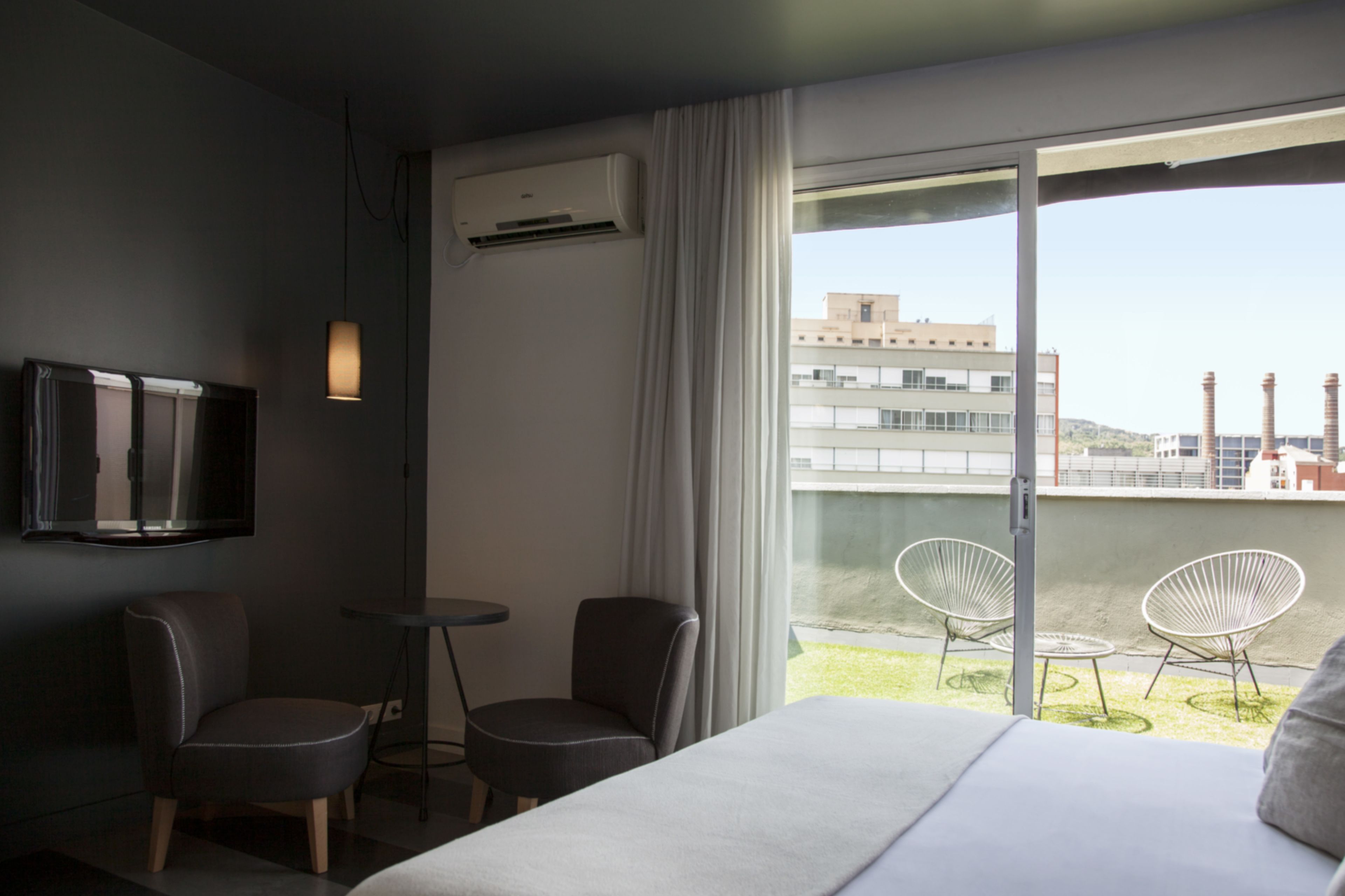 A hotel room with a bed, two chairs, a small table, a TV, and a balcony with chairs and a view of buildings.
