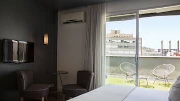 A hotel room with a bed, two chairs, a small table, a TV, and a balcony with chairs and a view of buildings.