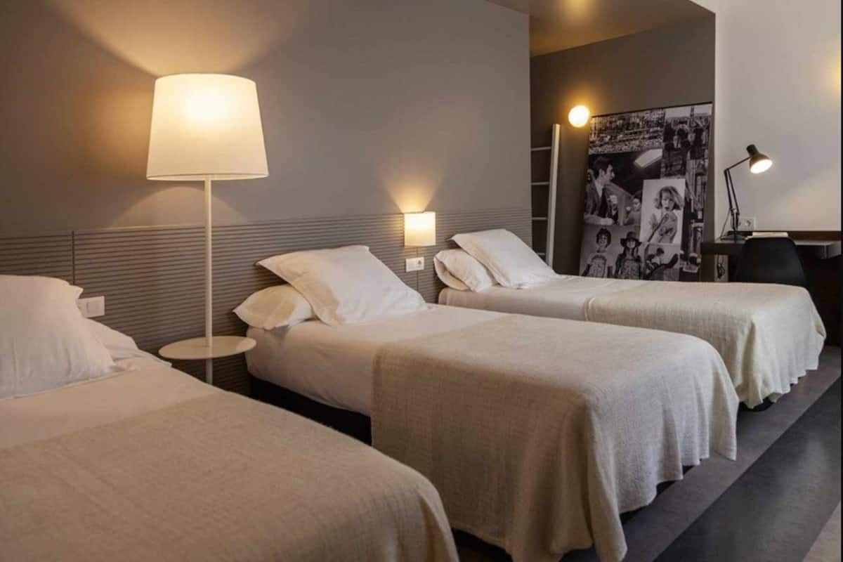 Una habitación de hotel con dos camas individuales, una mesita de noche con lámpara, un estante en la pared y un cartel.