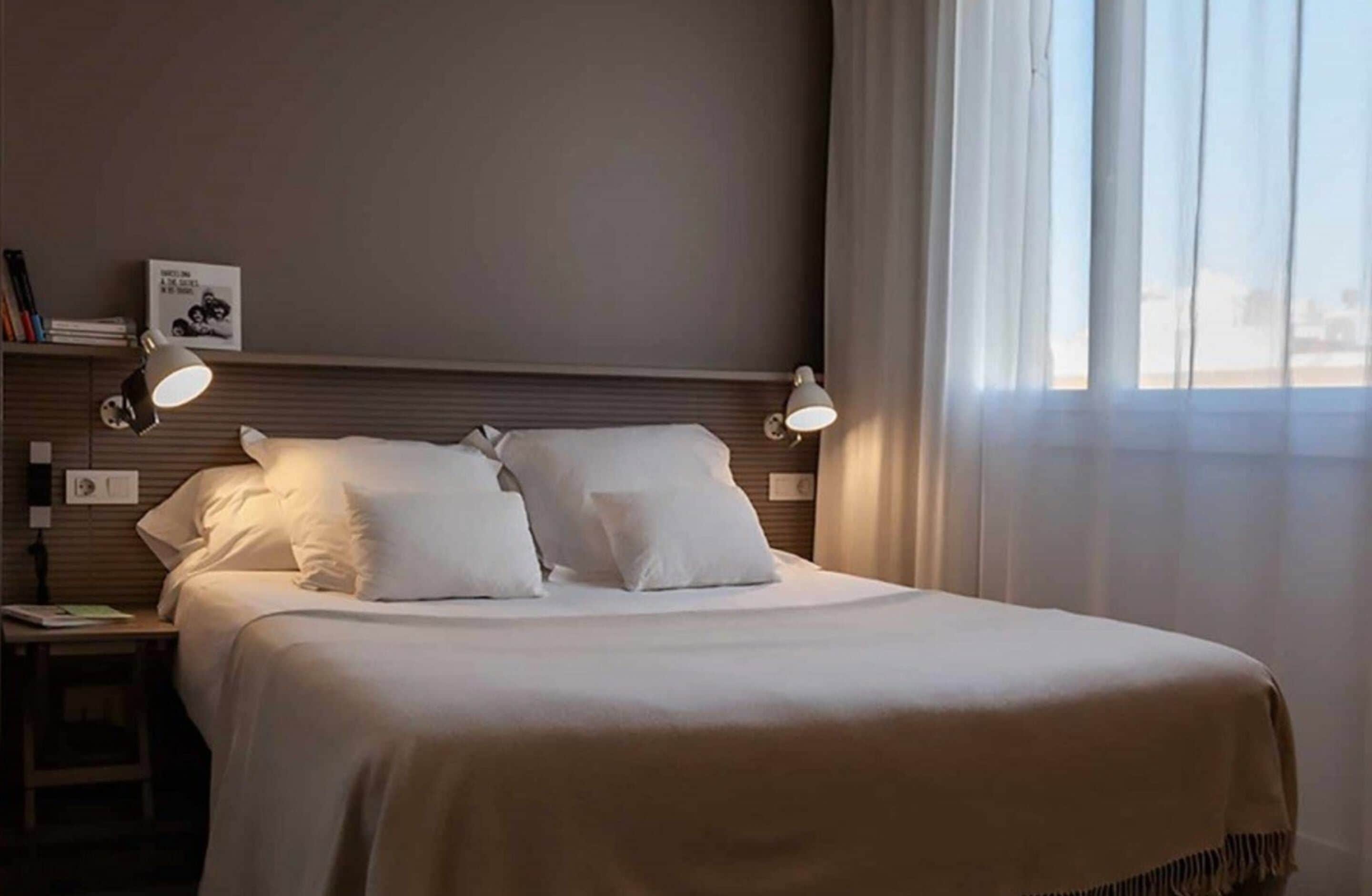 Una cama bien hecha con sábanas blancas, dos lámparas de noche, una mesita de noche con lámpara, un cuadro en la pared y una ventana con cortinas translúcidas.
