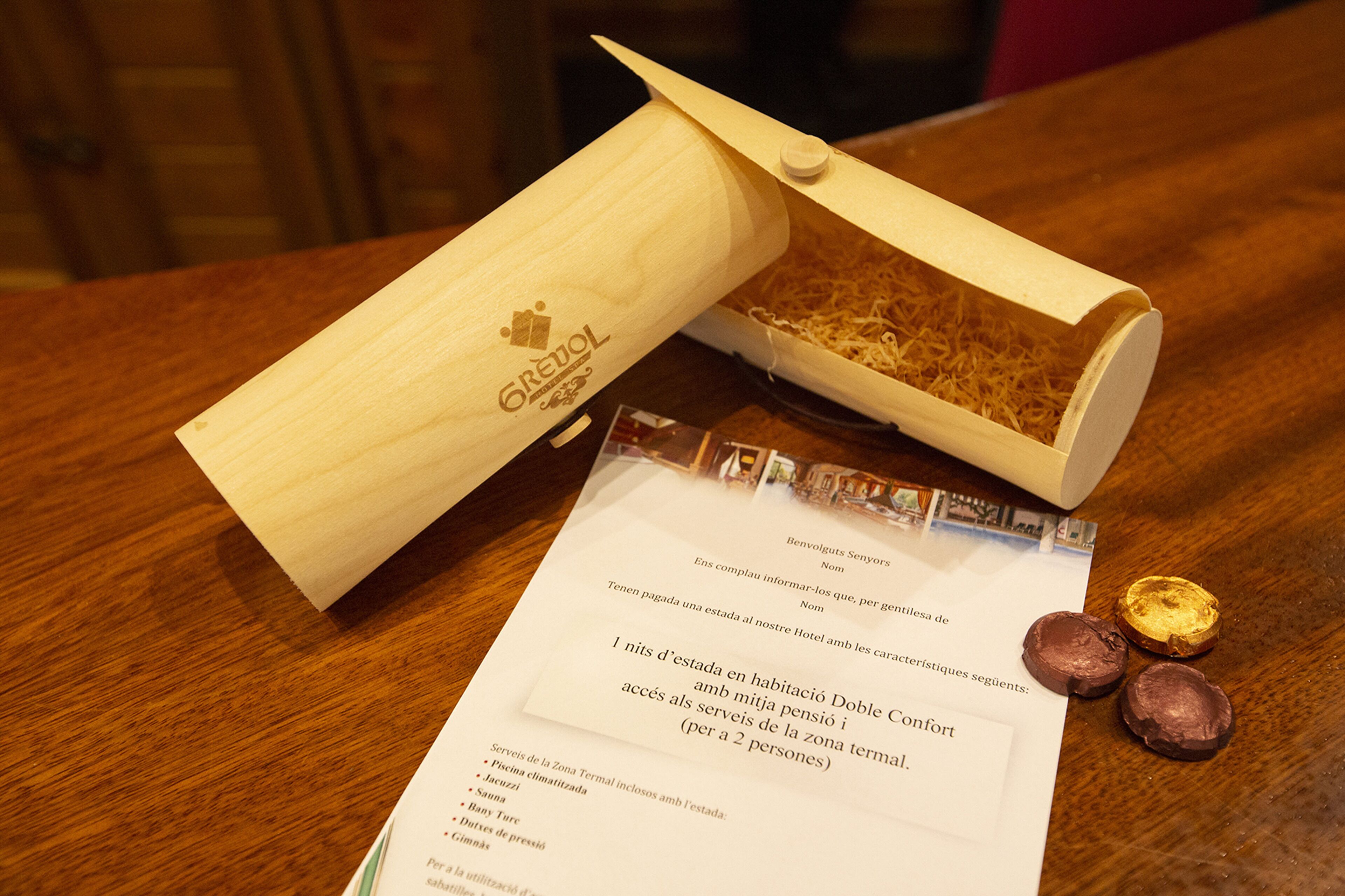 Una caja de madera con una etiqueta, un folleto y dos bombones sobre una mesa de madera.