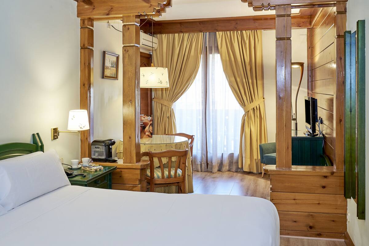 Ein Hotelzimmer mit einem Bett, einem Schreibtisch mit Lampe, einem Sessel, einem Fernseher und einem Fenster mit Vorhängen.