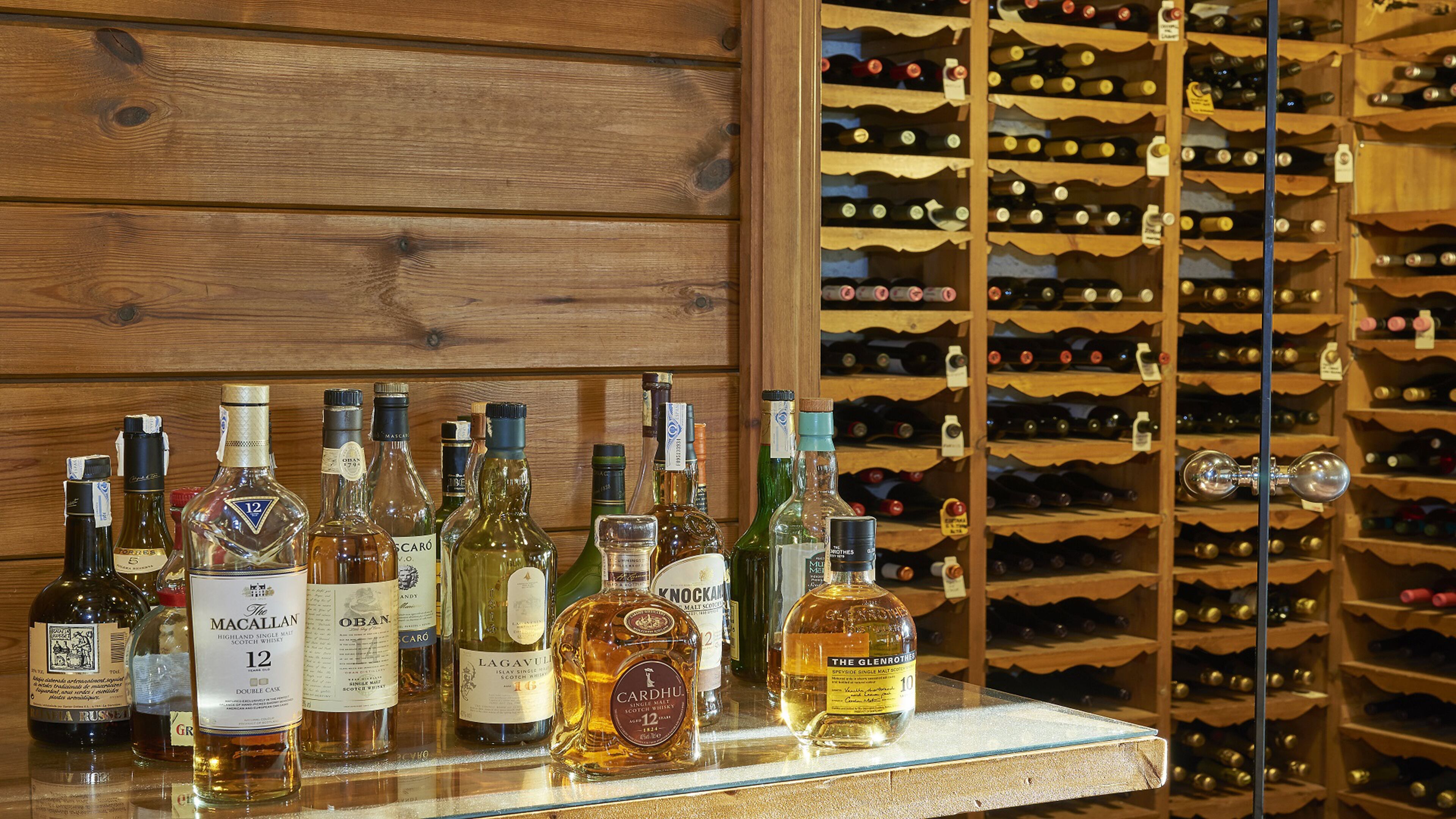 A variety of whisky bottles displayed on a shelf against a wooden wall, with a wine rack in the background.