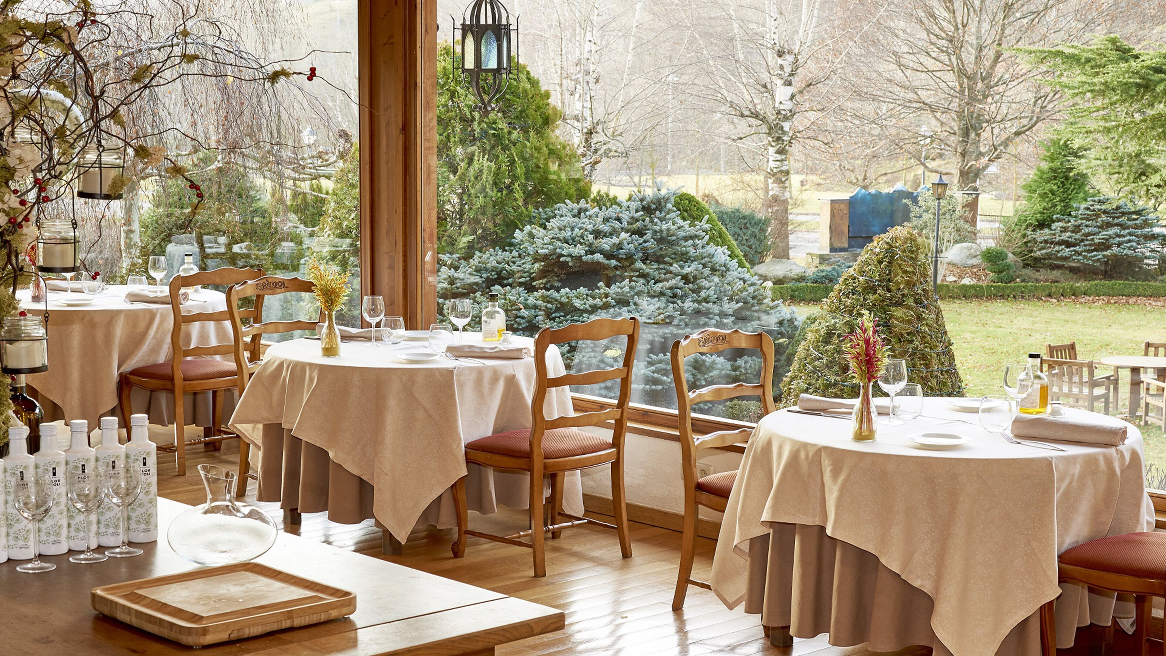 Un restaurante con mesas redondas, sillas de madera y vistas a un jardín con árboles, incluyendo un árbol de Navidad.