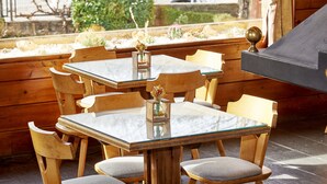 A wooden outdoor dining area with a glass table and chairs, a small vase with flowers, and a piano in the background.