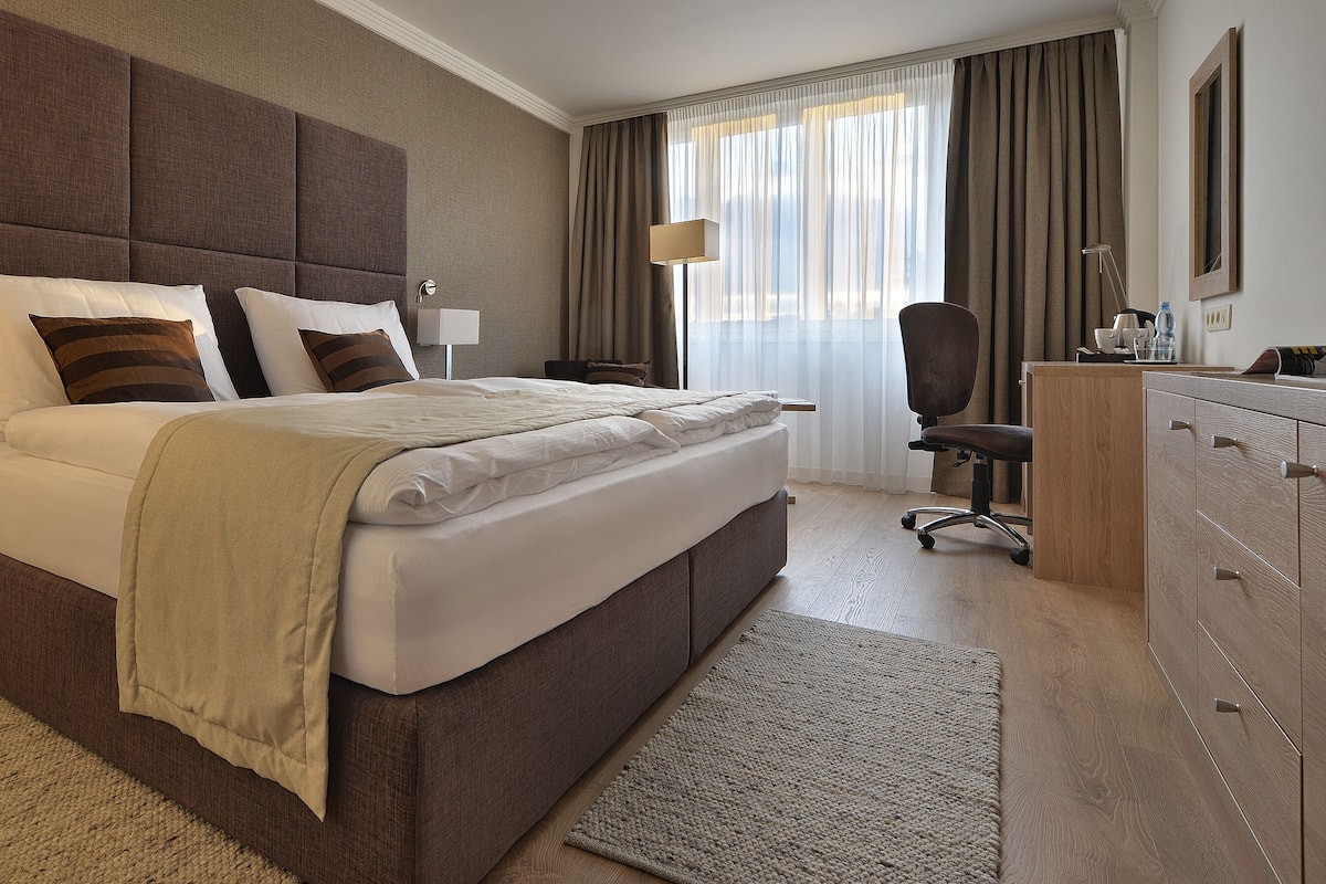A hotel room with a large bed, bedside tables, a desk, a chair, and a wardrobe.