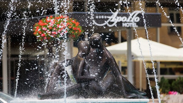 Une sculpture de fontaine avec des jets d’eau en mouvement, placée devant l’enseigne d’un hôtel et la façade d’un immeuble.