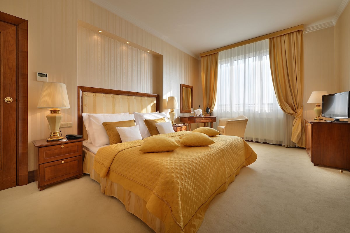 A hotel room with a large bed, a bedside table with a lamp, a dresser, a television, and a window with curtains.